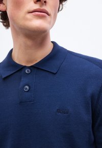 Άνδρας που φοράει μπλε ναυτικό πόλο πουκάμισο με δύο κουμπιά και μικρό κεντημένο λογότυπο "BOSS" στο στήθος.