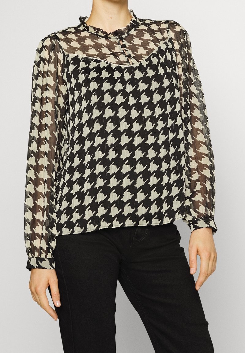 Femme portant un chemisier transparent à manches longues avec un motif pied-de-poule noir et blanc et un pantalon noir, debout devant un fond uni.