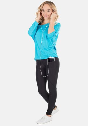 Turquoise langemouwtop, zwarte leggings met een telefoonvak, en witte sneakers. Model met oordopjes, met los en golvend haar.