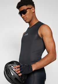 Schwarzes ärmelloses Sporttop mit strukturiertem Muster, hält einen schwarzen Fahrradhelm. Quadratische Sonnenbrille mit dunklen Gläsern.