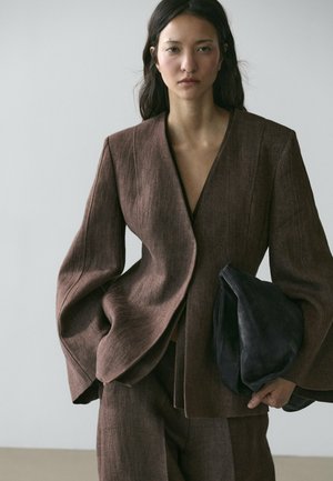 Frau in übergroßem braunem Anzug hält große schwarze Clutch, steht vor schlichtem hellem Hintergrund.