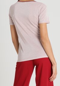 Lyserød kortærmet top med glitrende finish og rød kant ved halsen. Materialet ser glat ud; vist iført med røde bukser.