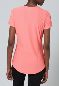 Persona con una camiseta ajustada de manga corta en color coral-rosado vista desde atrás, combinada con pantalones negros, sobre un fondo gris liso.