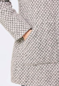 Manteau gris à motifs avec un tissu texturé à tissage en losange, une poche latérale visible et un détail de poignet légèrement retroussé.