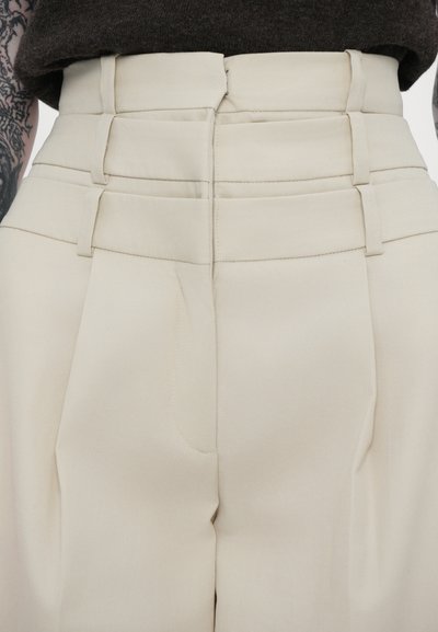 High-waisted beige pants made of smooth fabric, featuring folded details along the waistband and front pleats for structured shape.