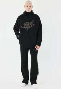Sort lynlås hoodie med et grafisk design i brun og matchende sorte bukser. Begge dele har en afslappet pasform og er lavet af blødt stof.