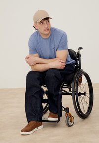 Young man wearing beige cap, blue t-shirt, dark pants, and brown sneakers sitting in a black wheelchair, looking to the side.