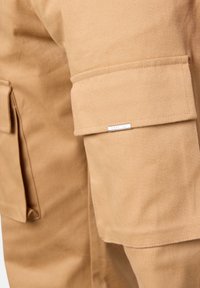 Khaki cargo pants made of durable fabric, featuring side pockets and a small metal logo accent. Smooth texture with clean stitching.