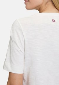 Weißes T-Shirt mit kurzen Ärmeln aus weichem, strukturiertem Stoff. Verfügt über ein dekoratives, gesticktes Logo auf der Rückseite in der Nähe des Halsausschnitts.