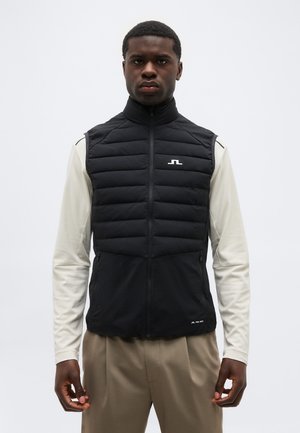 Mož močen v črni prešiti brezrokavnik čez belo srajco z dolgimi rokavi in bež hlačami, stoji pred enobarvnim ozadjem.