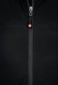 Veste softshell noire avec une fermeture éclair centrale dotée d'un tirette rouge proéminent et des coutures plates, présentant une texture lisse et mate.