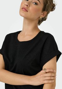 Sort kortærmet bluse med rund halsudskæring, der har en subtil glans. Gyldne hoopsøreringe og en ring med dekorativ detalje på hånden.