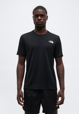 Joven de pie mirando hacia adelante, vestido con una camiseta negra de The North Face y pantalones cortos negros a juego, sobre un fondo claro y sencillo.