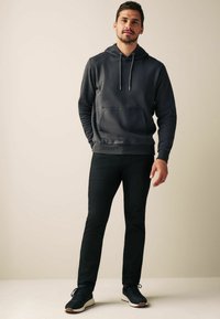 Sweatshirt à capuche gris foncé avec cordons de serrage, poche avant, pantalon slim noir, et baskets bleu marin avec semelles claires, se tenant devant un fond neutre.