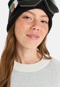 Knitted black beanie with a green and gray accent; model wearing a light gray sweater with a ribbed neckline; smooth skin with freckles.