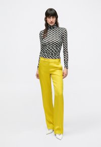 Top de cuello alto con patrón en blanco y negro y mangas largas, combinado con pantalones anchos de tiro alto en amarillo brillante y zapatos blancos puntiagudos.