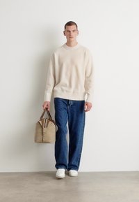 Beige strikkegenser, blå vide jeans, hvite sneakers, holder en beige veske med mørkebrune stropper og vertikale striper. Enkel design.