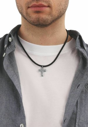 Hombre joven con ligera barba lleva un colgante de cruz plateada en un cordón negro sobre una camiseta blanca y una camisa gris abotonada abierta.