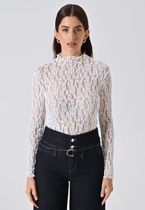 Top lungo bianco in pizzo con maniche a sbuffo e collo alto, caratterizzato da motivi floreali e una trama intricata, abbinato a jeans in denim scuro a vita alta.