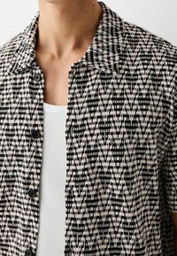 Camisa de manga corta con cuello y un patrón abstracto en negro y beige sobre una camiseta de tirantes blanca con estrías, usada por una persona.