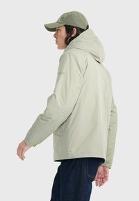 Hellgrüne Kapuzenjacke mit elastischen Bündchen, Fronttaschen und einer glatten Textur. Kombiniert mit dunkelblauen Jeans und einer passenden Mütze.