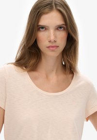 Lyserød t-shirt med rund hals i blødt stof, med korte ærmer og et diskret logo på venstre bryst. Glat tekstur.