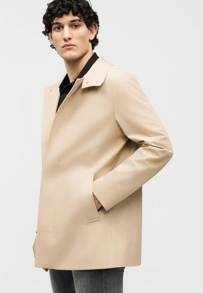 Ung mand iført beige trenchcoat over sort skjorte, med hånden i lommen, kigger mod kameraet foran en ensfarvet baggrund.