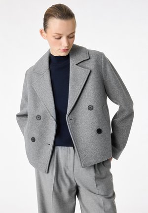 Kobieta ubrana w szarą wełnianą marynarkę dwurzędową, granatowy golf i szare dopasowane spodnie, patrząca w dół, z rękami w kieszeniach.