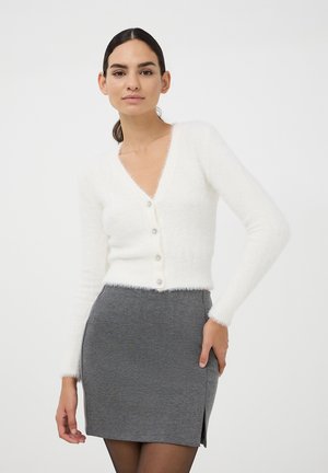 Cardigan bianco fluffy con scollo a V e cinque bottoni. Abbinato a una mini gonna grigia con uno spacco laterale. Tessuto morbido e stile aderente.