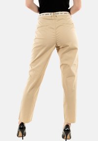 Beige high-waisted broek met een rechte pasvorm, voorzien van een witte elastische tailleband en twee achterzakken. Draag met zwarte hakken.