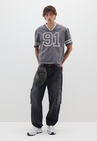 Jersey gris de manga corta con estampado blanco "91" y rayas, combinado con pantalones cargo negros, una gorra negra y zapatillas grises.