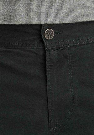 Pantaloni verde scuro con un bottone con la scritta "PME Legend Jeans" allacciato sotto la vita di un maglione grigio a coste.