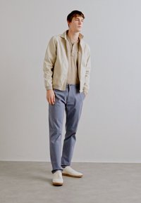Beige jakke, lysebrun polo og lyseblå bukser. Hvide sneakers med brune såler. Casual fit, glatte teksturer og neutrale farver.