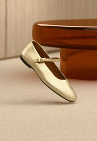 Goldene Ballerina aus Metall mit rundem Zehenbereich, Knöchelriemen und kleiner rechteckiger Schnalle, vor einem weichen, neutralen Hintergrund.