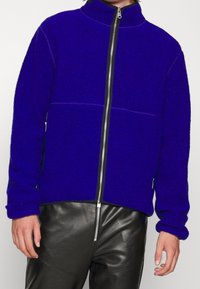 Polarowa kurtka zapinana na zamek w kolorze królewskim niebieskim, z wysokim kołnierzem, kontrastowymi zamkami błyskawicznymi i ściągaczami na rękawach. Połączona z czarnymi skórzanymi spodniami.