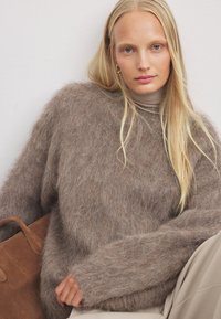 Brązowy, puszysty sweter dzianinowy z oversize'owymi rękawami, połączony z beżowym golfem i torebką ze skóry zamszowej w odcieniu brązowym. Teksturowany materiał i miękki wygląd.