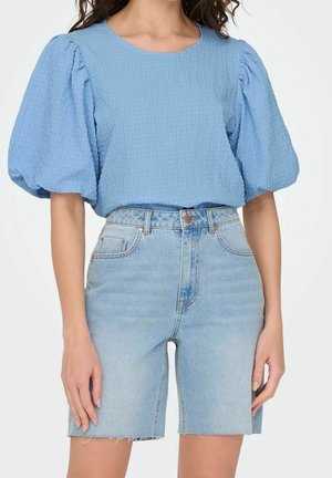 Femme portant un chemisier bleu clair texturé à manches bouffantes associé à un short en jean clair taille haute avec ourlets bruts.