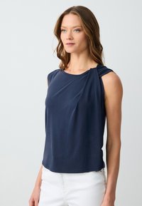 Top senza maniche blu navy con dettaglio arricciato sulla spalla sinistra; tessuto liscio con una vestibilità comoda, abbinato a pantaloni bianchi.