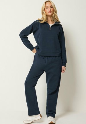 Mujer rubia con chándal azul marino con sudadera con cremallera hasta el cuarto y pantalones a juego, zapatillas blancas, de pie con una mano en la cadera.