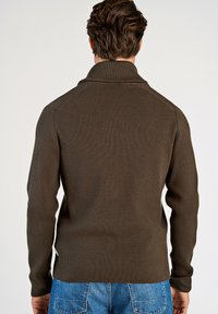 Braun gerippter Strickpullover mit hohem Kragen und langen Ärmeln, mit einem seitlichen Schlitz am Saum, kombiniert mit blauen Jeans.