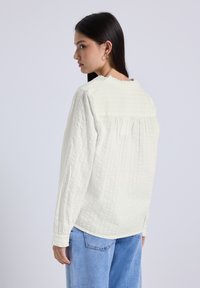 Blusa bianca leggera con maniche lunghe, scollatura arricciata e retro arricciato. Abbinata a jeans azzurri.