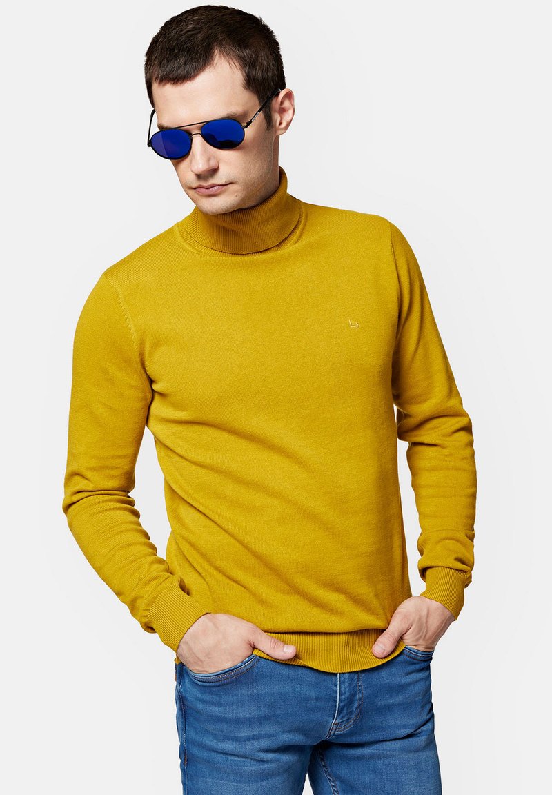 Musztardowozłoty sweter z kołnierzem typu turtleneck, wykonany z miękkiego materiału, z prążkowanymi mankietami i dołem. Noszony z niebieskimi okularami przeciwsłonecznymi i niebieskimi dżinsami.