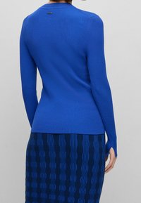 Pull bleu côtelé avec manches longues, col rond et un petit logo au dos. Associé à une jupe bleue texturée.