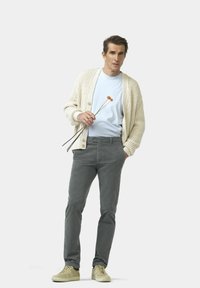 Strickjacke in Creme über einem hellblauen Hemd, kombiniert mit grauen Hosen und beigen Sneakern. Hält drei Stiele mit orangefarbenen Knospen.