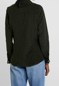 Mörkgrön kordfløyelskjorta med långa ärmar, med knappad manschett och rundad nederkant, visad bakifrån. Avslappnad passform.