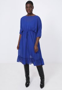Blauwe jurk tot op de knie met een losse pasvorm, een gathering in de taille en een gerimpelde zoom. Zacht, lichtgewicht materiaal en opgerolde mouwen. Draagt een paar zwarte laarzen tot de knie.