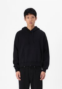 Sweatshirt preto com capuz, com um bolso frontal, design ligeiramente curto, punhos canelados e uma textura suave. Usado com calças pretas.