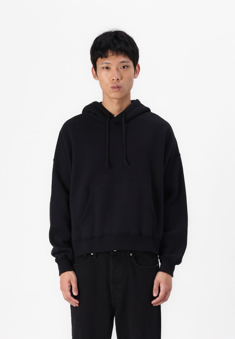 Sweatshirt preto com capuz, com um bolso frontal, design ligeiramente curto, punhos canelados e uma textura suave. Usado com calças pretas.