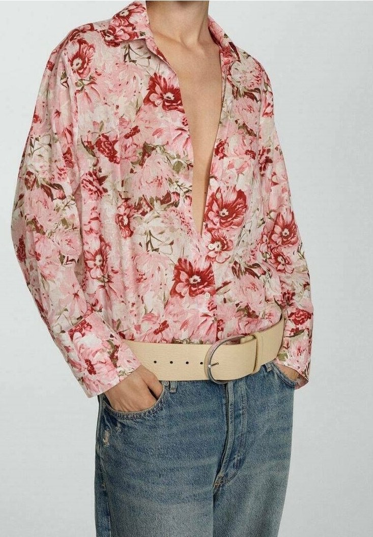 Personne portant un chemisier ample rose à fleurs avec un décolleté profond, rentré dans un jean bleu taille haute avec une large ceinture beige, les mains dans les poches.