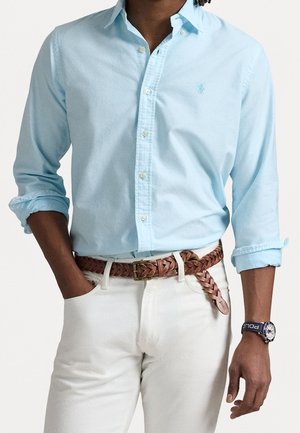 Personne portant une chemise bleu clair à boutons avec manches retroussées, pantalon blanc, ceinture tressée marron et montre bleu marine avec bracelet "Polo".